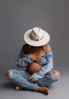 a pregnant woman in ripped jeans and a white hat is sitting on the floor with her arms around her stomach