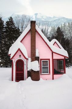 a small pink house with snow on the roof and windows, in front of some trees