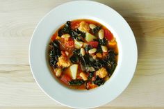 a white bowl filled with soup and vegetables on top of a wooden table next to a spoon