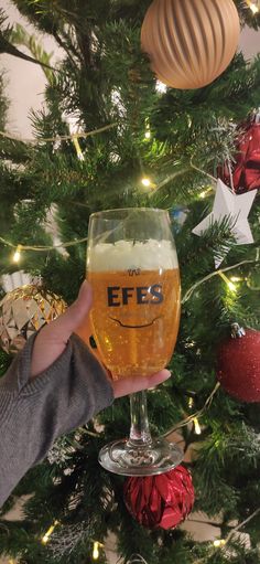 a person holding up a glass of beer in front of a christmas tree with ornaments