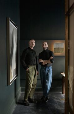 two men standing next to each other in an empty room with paintings on the wall