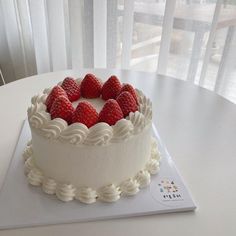 there is a cake with white frosting and strawberries on the top, sitting on a table next to a window