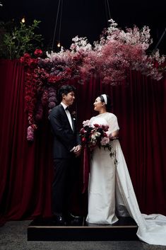 a man and woman standing next to each other in front of a red curtain with flowers