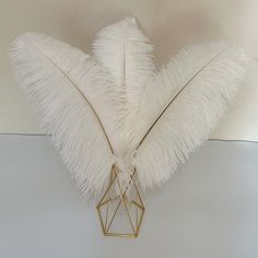 a white feather arrangement on a table with a gold geometric vase in the middle and two feathers sticking out of it