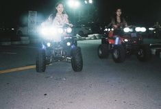 two people on four wheelers driving down the street at night with lights on them