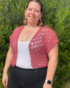 a woman wearing black pants and a pink cardigan smiles at the camera while standing in front of bushes