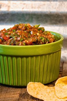 a green bowl filled with food next to some tortilla chips on top of a wooden table