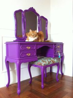 an orange cat sitting on top of a purple vanity with mirror and footstool