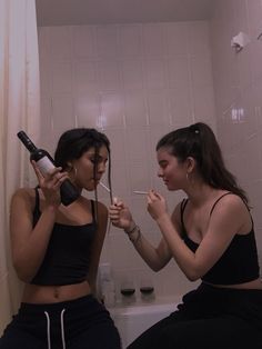 two young women sitting on a toilet brushing their teeth and holding wine bottles in each other's mouths