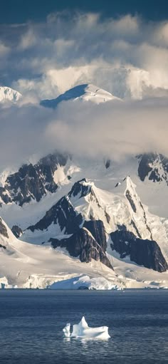 an iceberg floating in the ocean with snow covered mountains behind it and cloudy skies