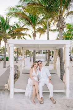 a man and woman sitting on top of a bed under palm trees at the beach