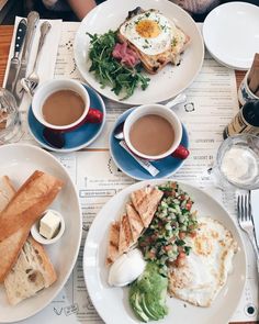 two plates with food on them sitting on a table next to cups of coffee and spoons