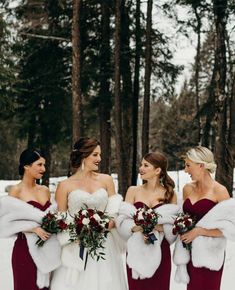 three bridesmaids in red dresses and fur stoles smile at each other while standing in the snow