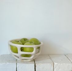 a white bowl filled with green apples sitting on top of a wooden block wall next to a white brick wall