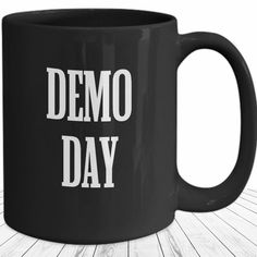 a black coffee mug with the words demo day on it sitting on a wooden table