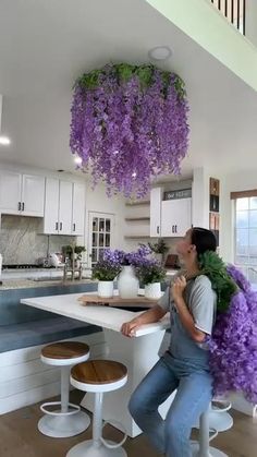 a woman sitting at a kitchen counter with purple flowers hanging from the ceiling