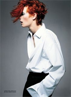 a woman with red hair wearing a white shirt and black pants, standing in front of a gray background