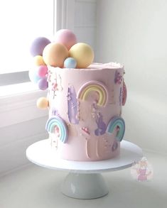 there is a pink cake with rainbows and balloons on the top, sitting in front of a window