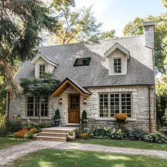 a stone house with white trim and two story windows, surrounded by greenery and flowers