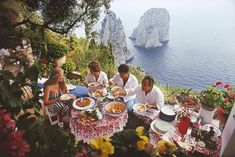 a group of people sitting around a table with food on it near the ocean and cliffs