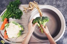 a person washing broccoli in a sink with carrots and cauliflower