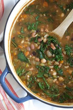 a pot filled with beans and greens on top of a white cloth next to a wooden spoon