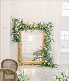 a mirror with flowers and greenery in front of it on a table next to chairs