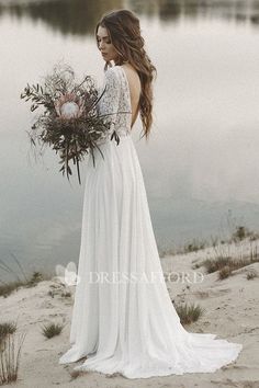 a woman in a white dress holding a bouquet of flowers standing on the shore of a lake