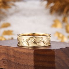 a gold wedding band with leaves on it sitting on top of a wooden block in front of a wall