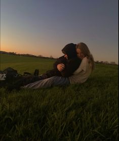 a man and woman sitting in the grass with their arms around each other as the sun sets