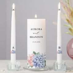 three white candles sitting next to each other on top of a table with pink and blue flowers