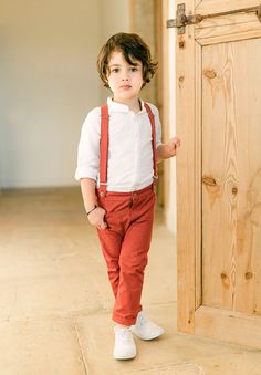 a little boy standing in front of a wooden door wearing red suspenders and a white shirt