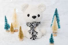 a crocheted white teddy bear sitting in the snow next to small christmas trees