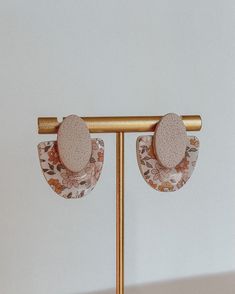 two pairs of earrings are hanging on a gold stand against a white wall with floral fabric