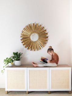 a woman reading a book on top of a white cabinet next to a gold sun mirror