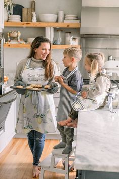 a woman and two children standing in a kitchen with cookies on the counter, smiling at each other