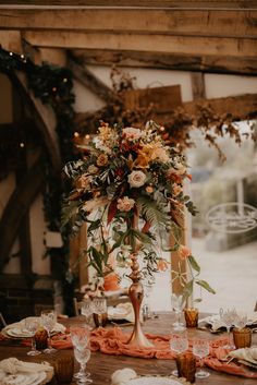 a tall vase with flowers and greenery on top of a wooden table surrounded by place settings