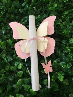 a white candle with pink and yellow butterflies on it sitting in front of some green bushes