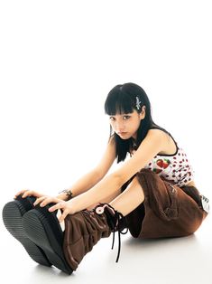 a young woman sitting on the ground with her legs crossed, wearing brown pants and boots