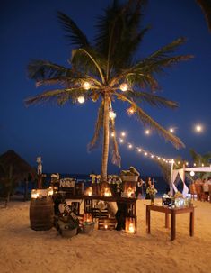 an outdoor dining area on the beach at night with lights strung from palm trees and hanging lanterns