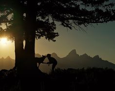 two people standing next to a tree with the sun setting behind them and mountains in the background