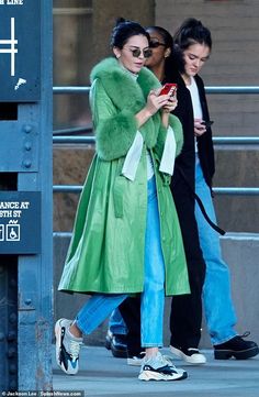 two women are walking down the street while looking at their cell phones and one is wearing a green coat