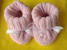 two pink knitted baby booties with white bows on the top and bottom, sitting on a yellow surface