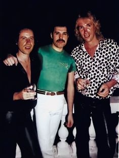 three men standing next to each other in front of a black background, one wearing a green shirt and the other white pants