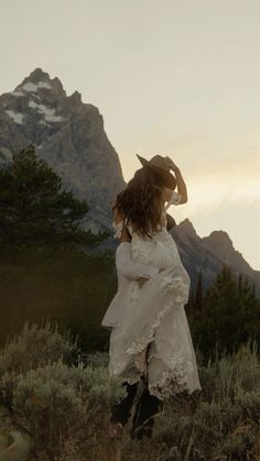 a woman wearing a white dress and cowboy hat standing in the grass with mountains behind her