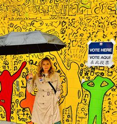 a woman holding an umbrella standing in front of a yellow wall with graffiti on it