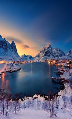 a lake surrounded by snow covered mountains with lights in the middle and houses on the other side