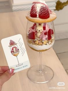 a person holding up a card next to a wine glass filled with dessert and strawberries