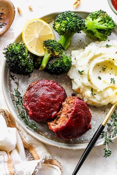 a white plate topped with meat, mashed potatoes and broccoli next to lemon wedges