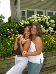 two young women standing next to each other in front of a flower garden and house